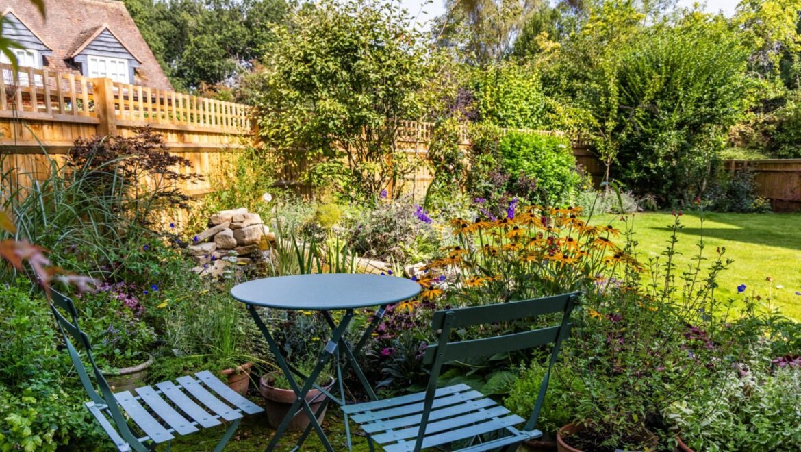Image of bistro garden set, surrounded by garden beds filled with flowering plants and a water feature